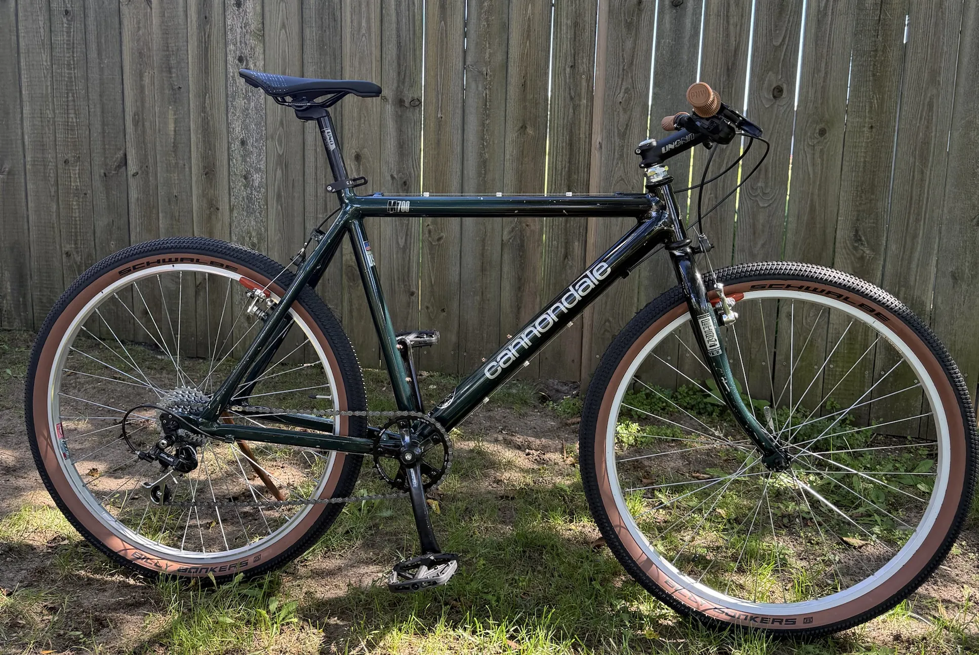 A dark green 1993 Cannondale M700 with tan-wall tires and tan grips, leaning in front of a weathered wooden fence.